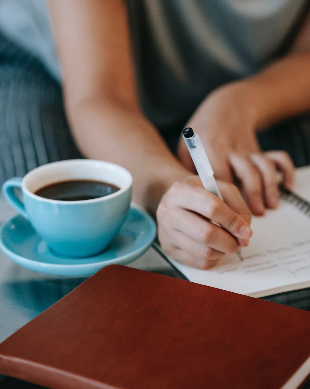 Woman writing in a notebook, with a book and a cup of coffee on the desk next to her